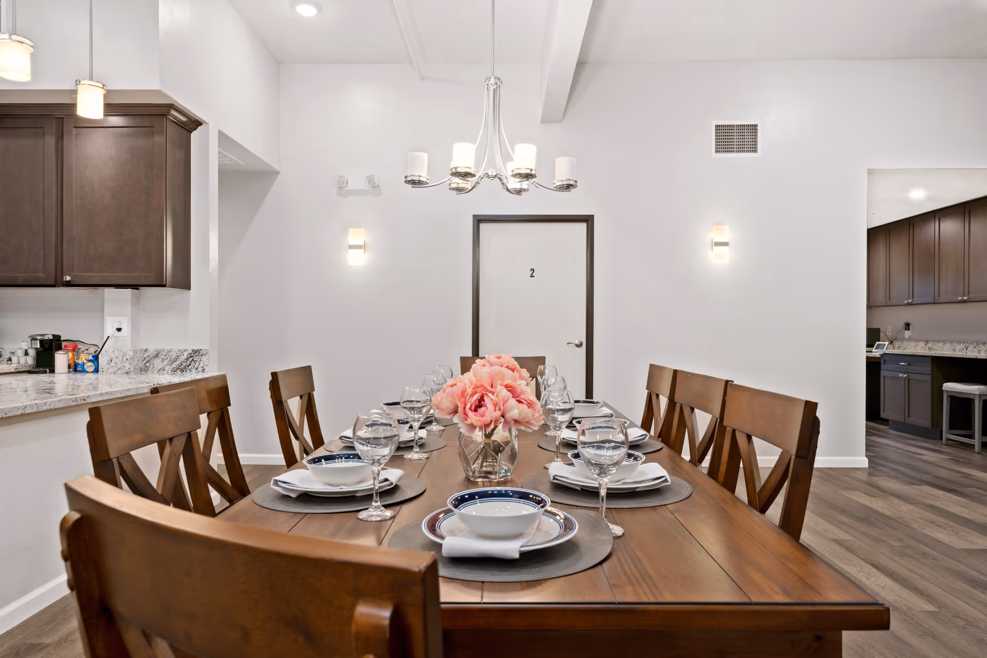 A dining room with a wooden table set with plates, glasses and a pink flower centerpiece, open to a kitchen area.