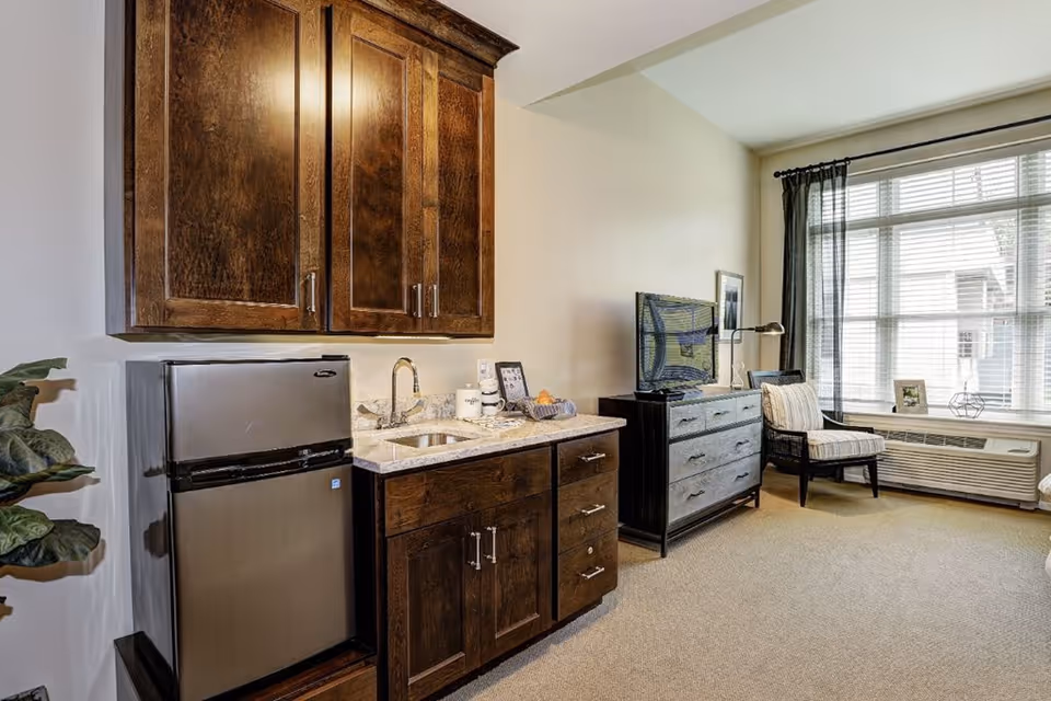 Interior view of a senior living facility room featuring a kitchenette with dark wooden cabinets, a small stainless steel refrigerator, and a granite countertop with a sink. Adjacent to the kitchenette is a living area with a dark wooden dresser, a flat-screen TV, a striped armchair, and a large window with blinds and sheer curtains allowing natural light to fill the room.