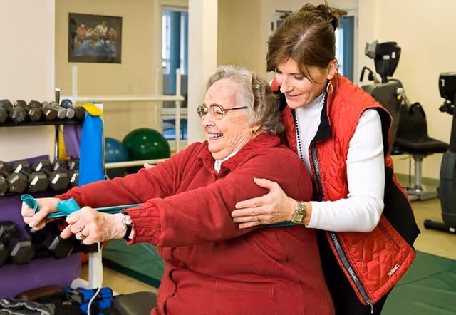 An elderly woman in a red sweater is exercising with a resistance band while a caregiver or trainer in a red vest assists and supports her in a fitness room with exercise equipment and weights in the background.