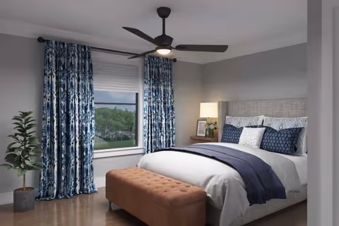 Bright bedroom with a upholstered bed dressed in blue and white linens, a ceiling fan, patterned curtains, a bench at the foot, and a potted plant by the window.