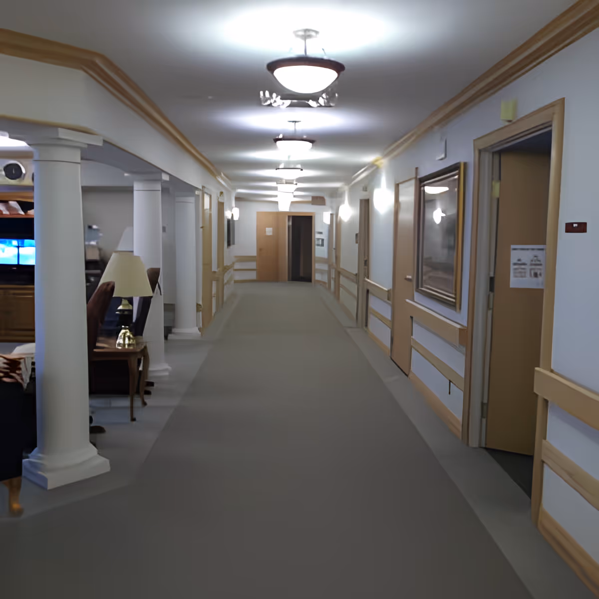 A long, well-lit hallway in a senior living facility with beige walls and carpeted floor. The hallway has several doors on the right side, a framed picture on the wall, and white columns on the left side leading to a common area with chairs and a lamp.