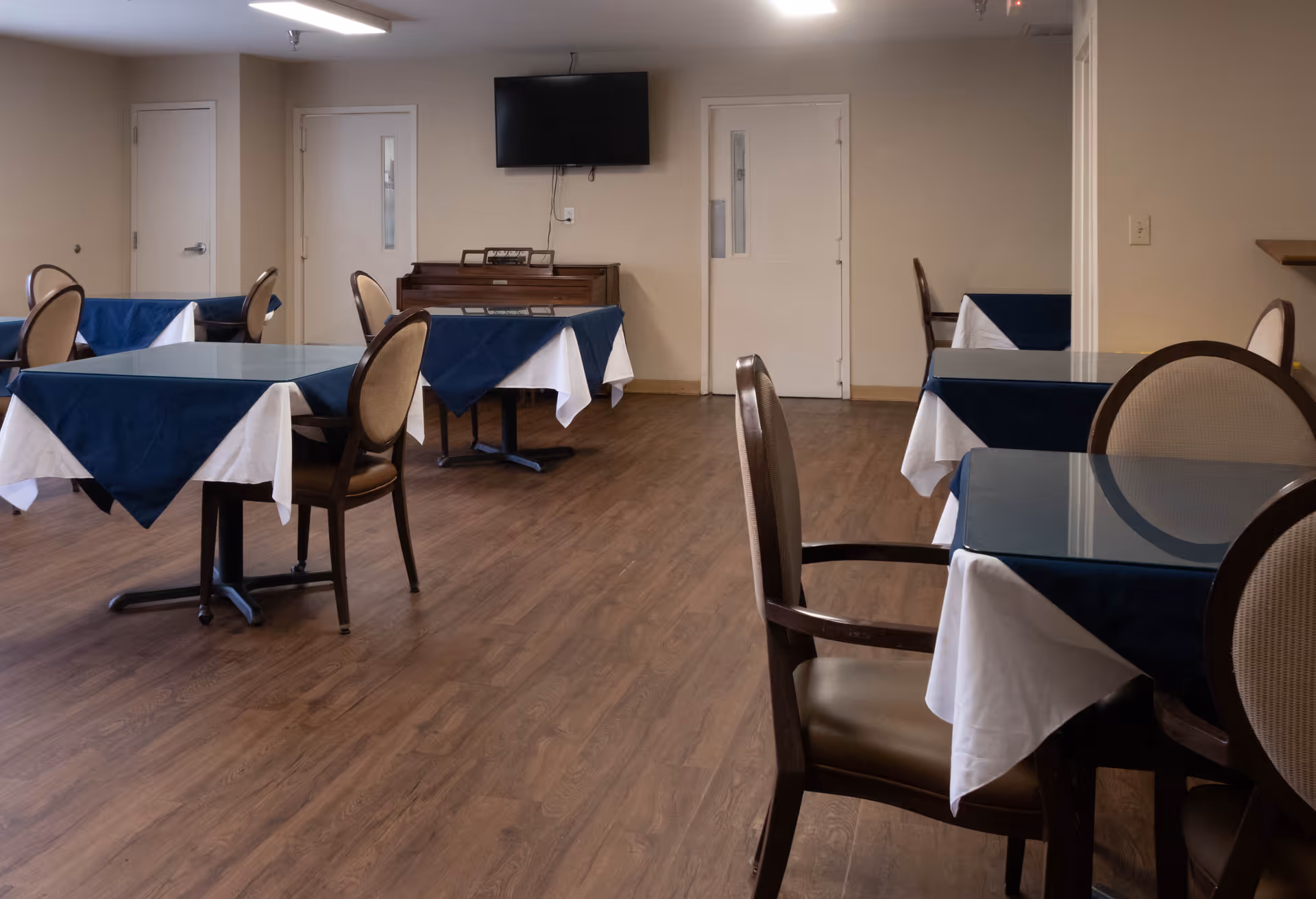 Empty dining room with tables draped in blue and white tablecloths, chairs, a piano and a wall-mounted TV.