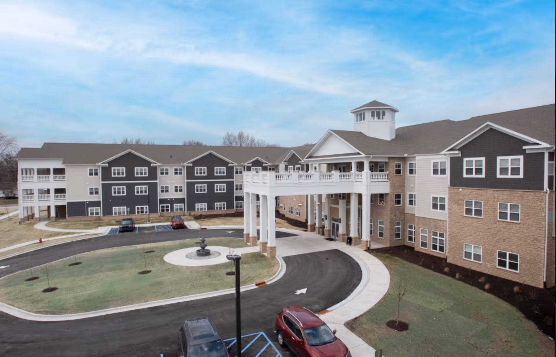 Exterior view of Avalon Senior Living facility showing a large, multi-story building with a covered entrance supported by white columns, a circular driveway with a small fountain in the center, several parked cars, and a partly cloudy blue sky.