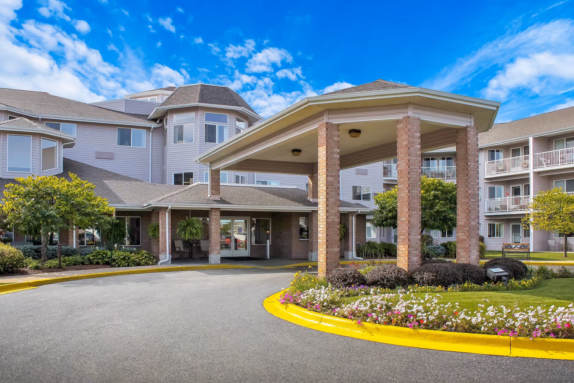 Exterior view of Independence Village of Saginaw (Wescourt) showing the main entrance with a covered drop-off area supported by brick columns, surrounded by landscaped flower beds and greenery under a partly cloudy blue sky.