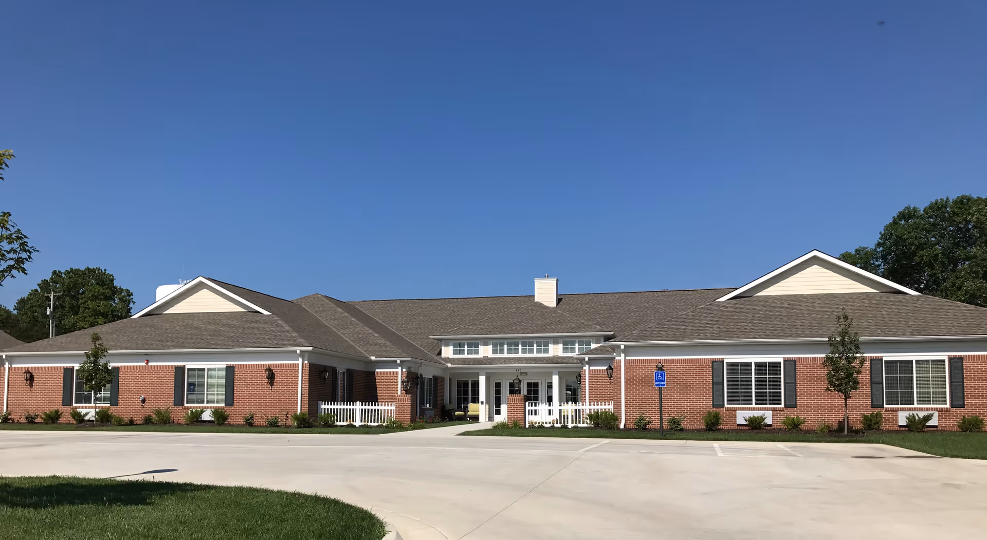 Single-story brick senior living building with a central covered entrance, white trim, and a parking area under a clear blue sky.