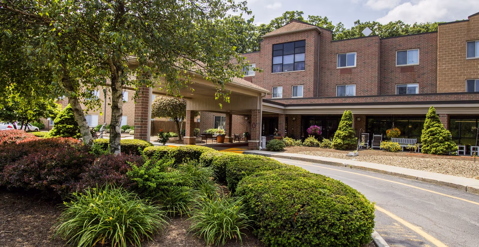 Front entrance of a brick senior living building with a covered drop-off, landscaped shrubs, and a driveway.