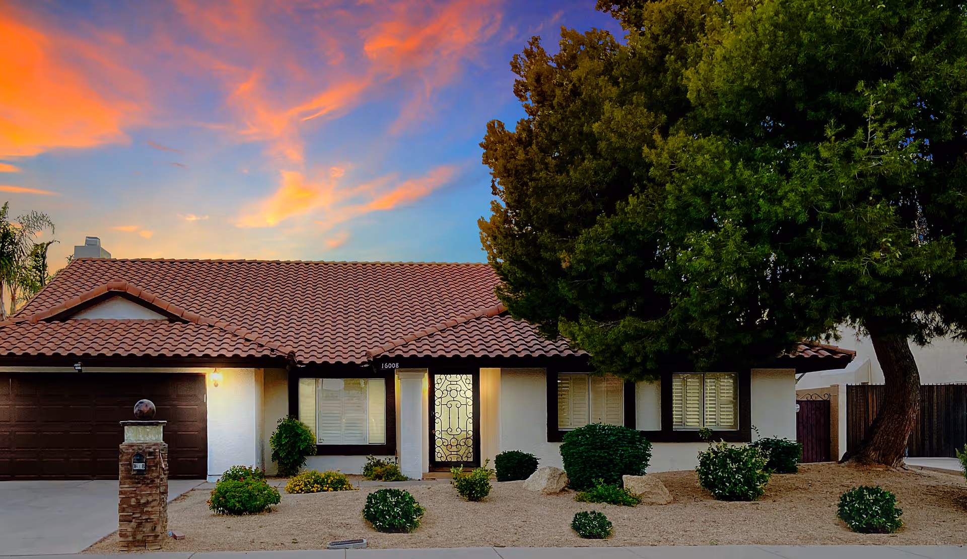 Front exterior of a single-story home with a red tile roof, garage, desert landscaping and a large tree at sunset.