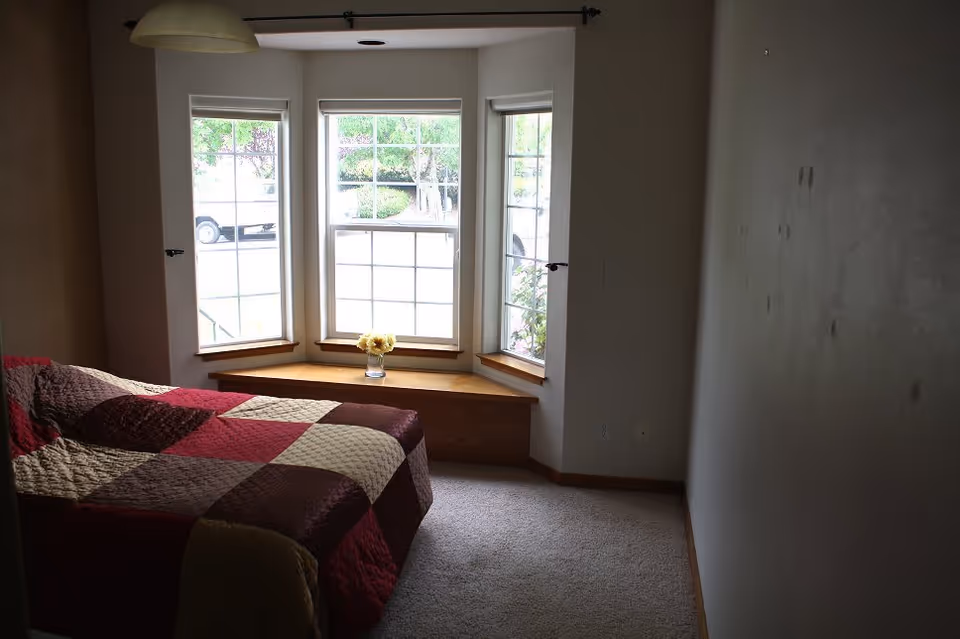 A bedroom with a bed covered in a patchwork quilt in shades of red, brown, and beige. The room features a bay window with three sections, letting in natural light. A small vase with yellow flowers is placed on the wooden window seat beneath the windows. The walls are plain, and the floor is carpeted.