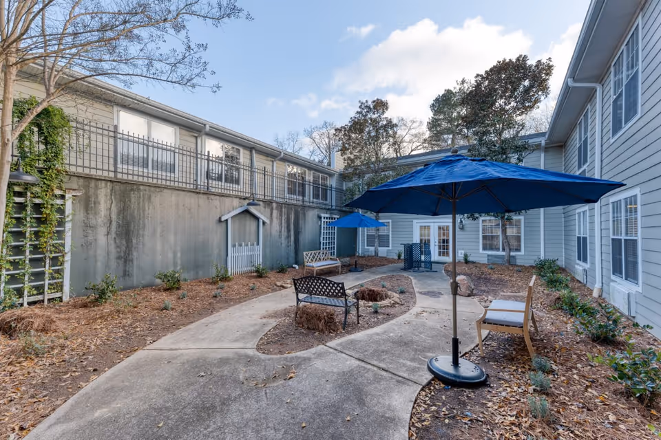 Outdoor courtyard area at Addington Place of Roswell featuring a concrete pathway, benches, blue umbrellas, and surrounding shrubs and trees with a two-story building in the background.
