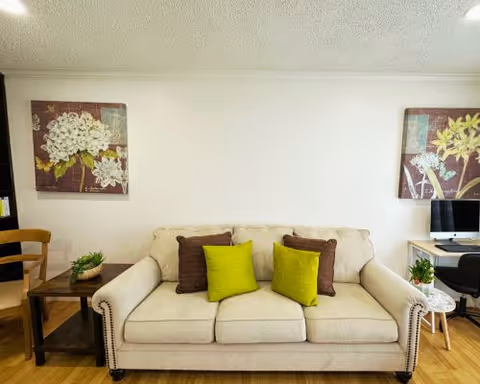 A cozy living room area with a beige sofa adorned with two green and two brown cushions. On either side of the sofa are small tables with plants and decorative items. The wall behind the sofa features two floral paintings. To the right, there is a desk with a computer and a black office chair.