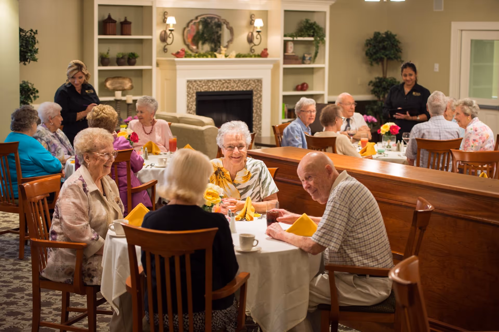 A group of elderly people sitting at round tables in a dining room, engaged in conversation and enjoying drinks. Two staff members are standing nearby, taking orders or assisting. The room has warm lighting, wooden chairs, white tablecloths, yellow napkins, and a fireplace with decorative shelves in the background.