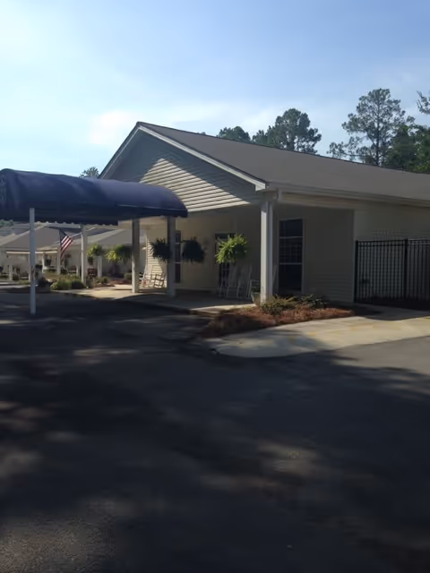 Exterior view of a single-story building with a covered entrance featuring a blue awning. There are hanging plants and rocking chairs on the porch area. Trees are visible in the background under a clear sky.