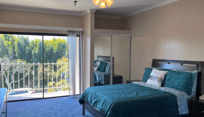 A bright bedroom with a large sliding glass door leading to a balcony with green trees outside. The room features a bed with teal bedding and pillows, a mirrored closet door, and a nightstand with a book on it. The walls are painted beige and the floor is carpeted in blue.