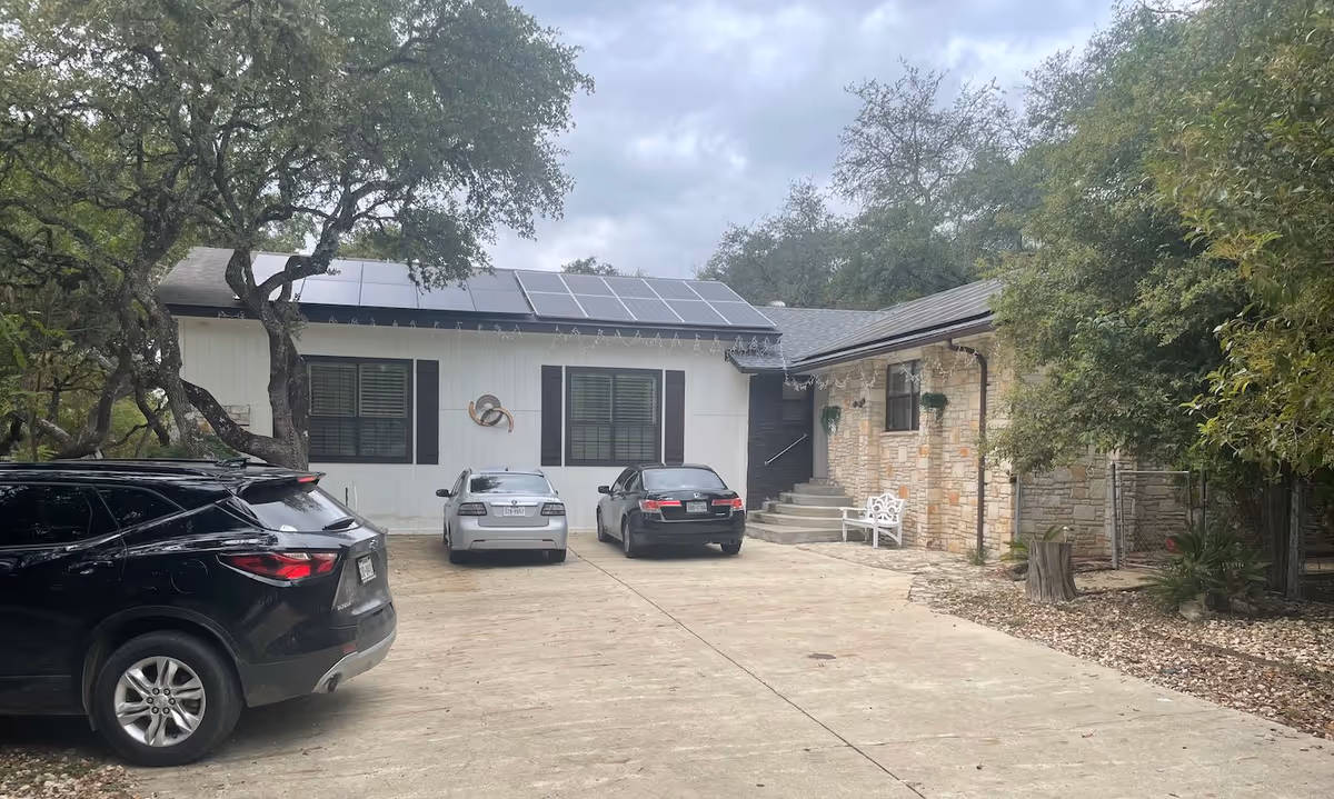 Exterior view of a single-story assisted living facility building with a stone and white panel facade, solar panels on the roof, three parked cars in the driveway, and trees surrounding the area under a cloudy sky.