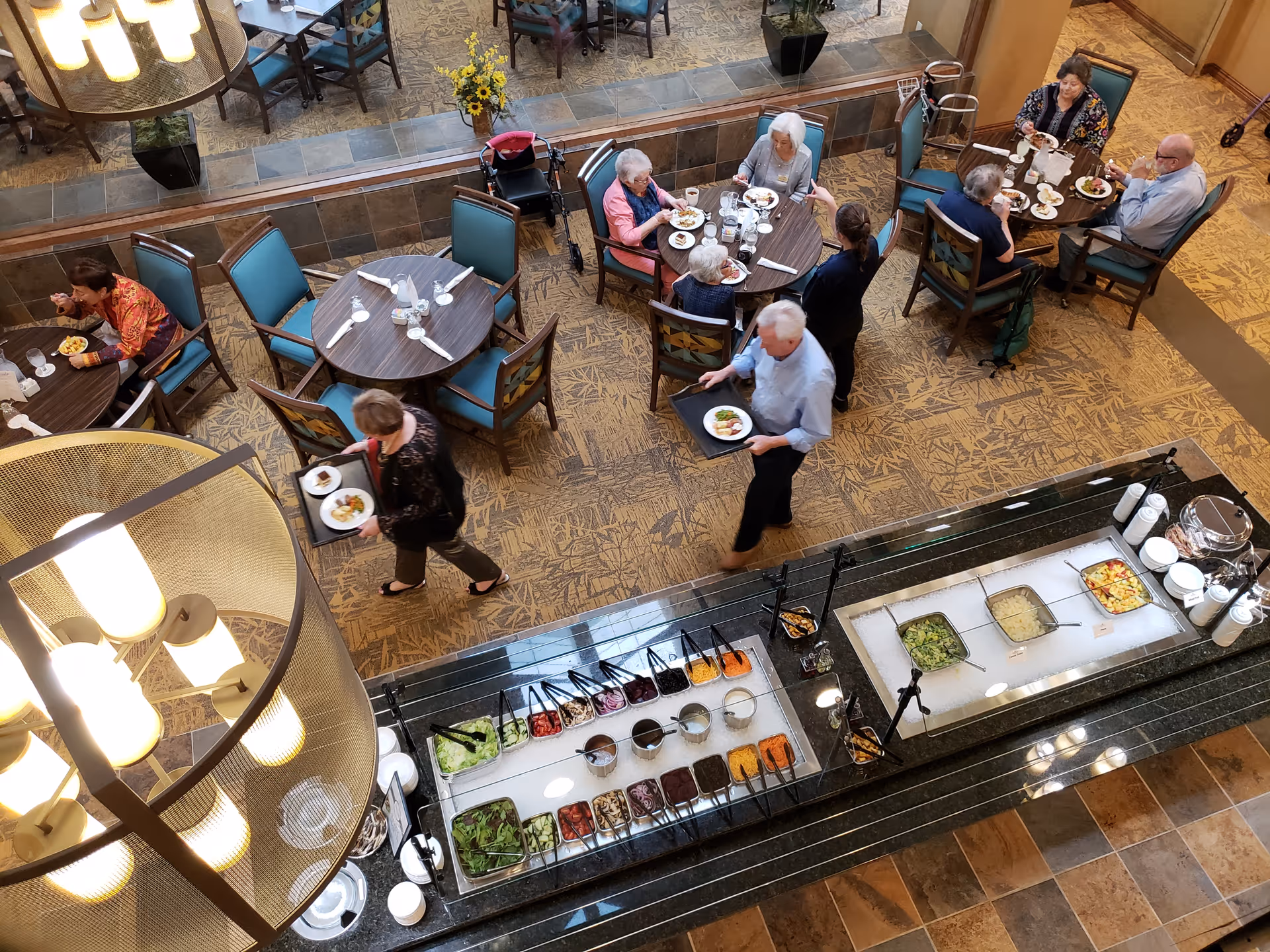 Overhead view of a dining area in a retirement community with several elderly people seated at round tables eating meals. A salad bar with various fresh vegetables and toppings is visible in the foreground. Two staff members are serving or assisting residents.