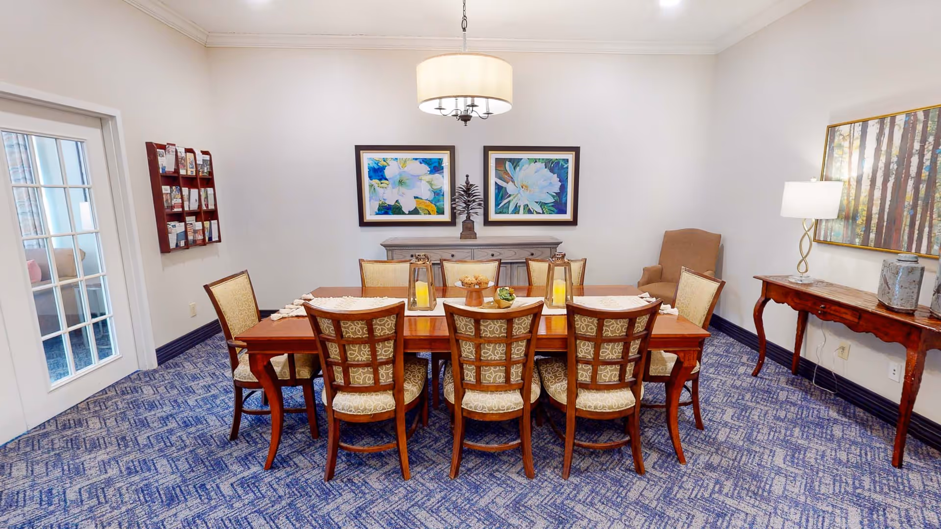 Dining room with a wooden table set with lanterns and eight upholstered chairs beneath a hanging light, framed artwork on the walls, and patterned blue carpet.