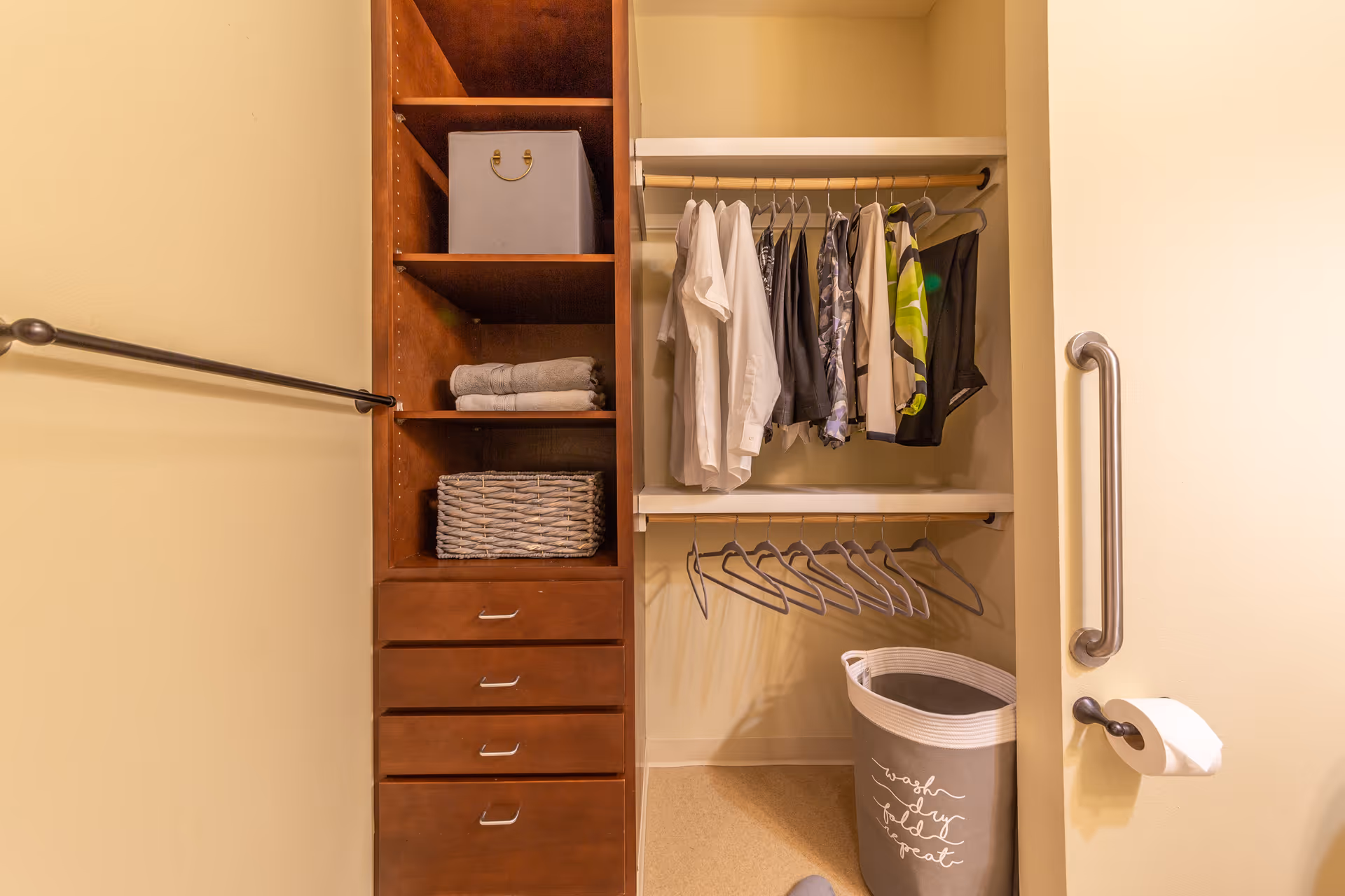 Open closet with wooden shelving and drawers, hanging clothes on a rod and a laundry basket, with a grab bar and toilet paper holder visible on the right.