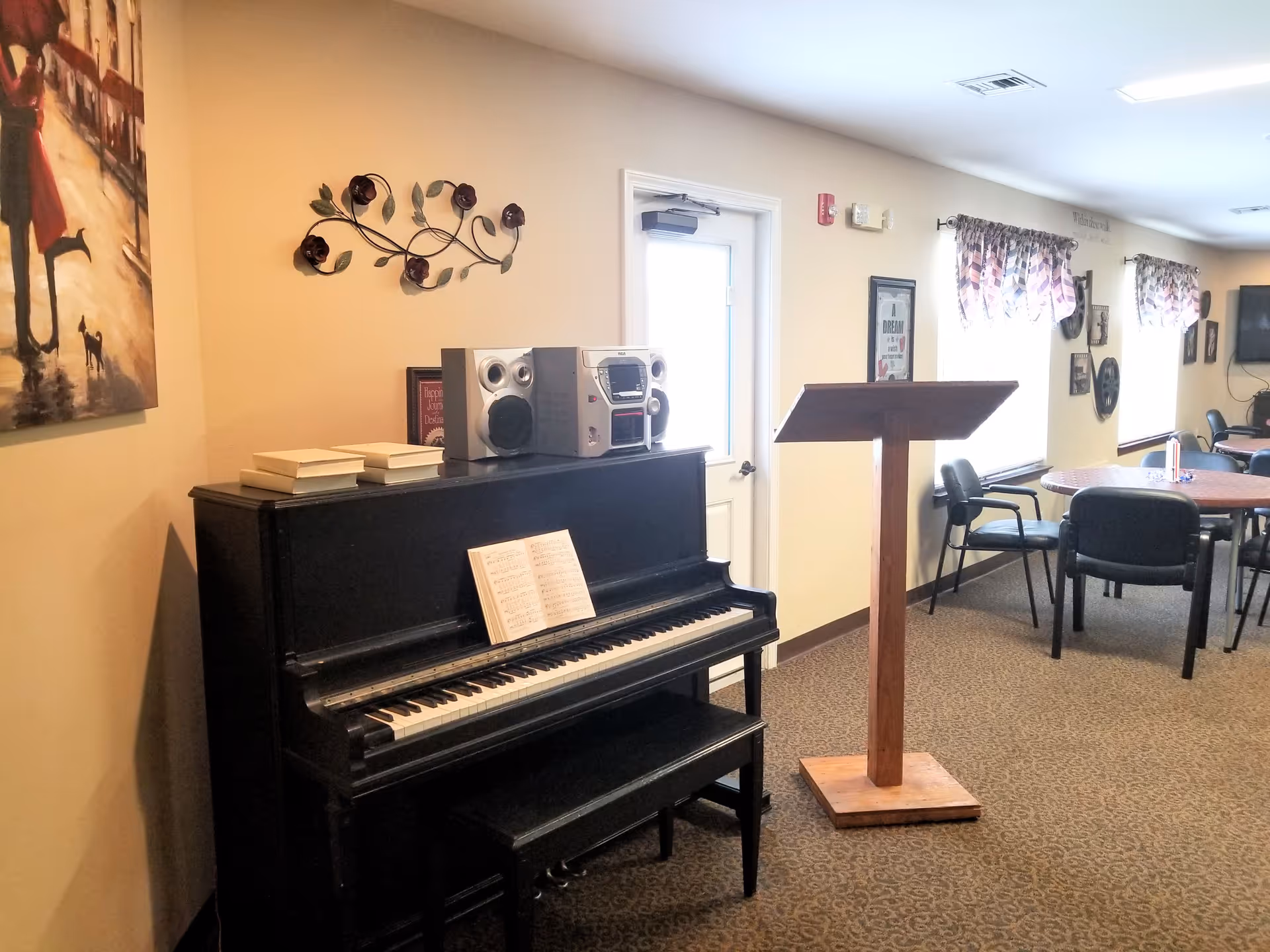 Interior view of a common area in a senior living facility featuring a black upright piano with sheet music and a stereo on top, a wooden music stand, round tables with chairs, wall decorations, and windows with patterned curtains.