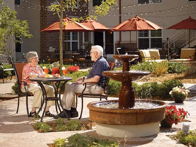 An elderly man and woman sitting at a round outdoor table with drinks, enjoying a sunny day in a garden courtyard with a three-tiered water fountain in the foreground and red umbrellas providing shade in the background.