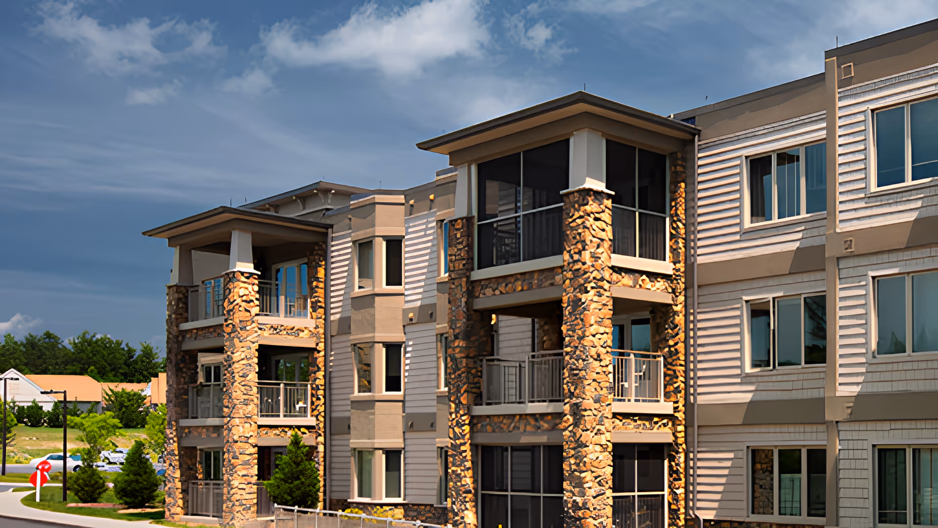 Three-story stone-and-siding senior living building with balconies and landscaped grounds under a partly cloudy sky.