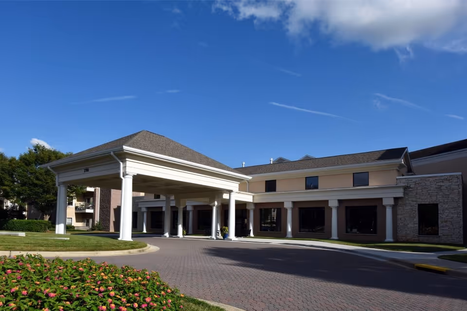 Exterior view of The Forest at Duke senior living facility showing a covered entrance with white columns, a paved driveway, landscaped greenery, and a clear blue sky.