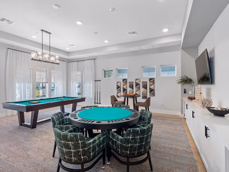 A bright and spacious game room featuring a pool table with cues and balls, a round card table with green felt surrounded by four green upholstered chairs, and a small table with two chairs against the wall. The room has large windows with sheer white curtains, modern light fixtures, and a mounted flat-screen TV above a white cabinet with decorative items.