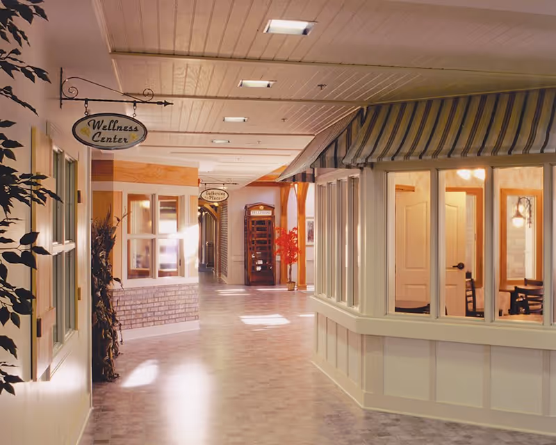 Indoor hallway of a senior living facility with signs for a Wellness Center and Gathering Place. The hallway has a light-colored tiled floor, white paneled walls, and a ceiling with recessed lighting. There is a vintage-style red telephone booth at the end of the hallway and a windowed room with striped awnings on the right side. Potted plants and warm lighting create a welcoming atmosphere.