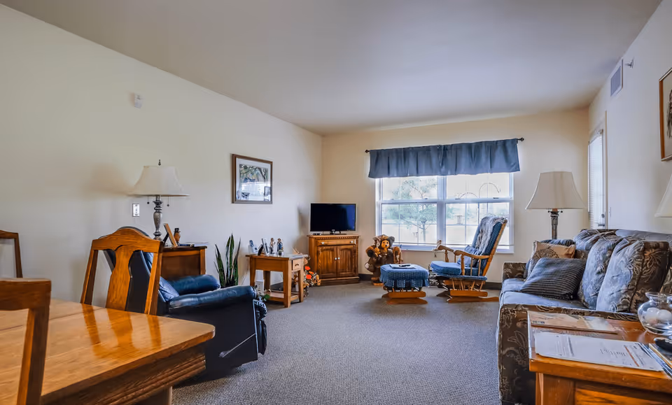 A cozy living room in a retirement village featuring a wooden dining table and chairs, a blue recliner, a wooden TV stand with a flat-screen TV, a rocking chair with a matching ottoman, a sofa with patterned cushions, two table lamps, a large window with a blue valance, and various decorative items including framed pictures and plants.