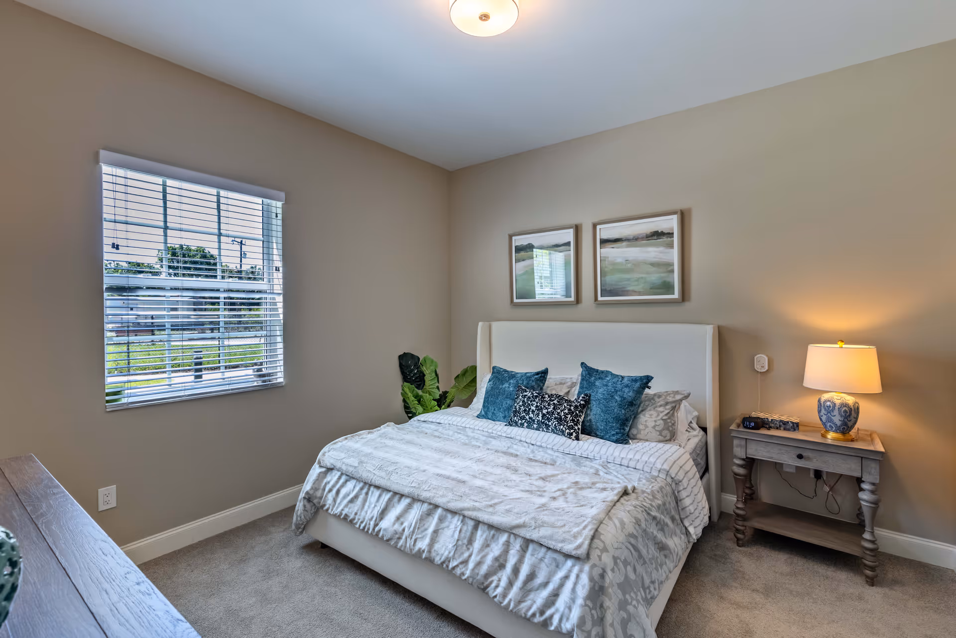 A cozy bedroom with a large bed featuring white and patterned bedding and multiple decorative pillows. There is a wooden nightstand with a blue and white lamp on the right side of the bed. Two framed landscape paintings hang on the beige wall above the bed. A window with white blinds is on the left wall, letting in natural light. The room has beige walls and carpeted flooring.