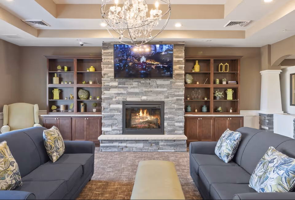 A communal living room with a stone fireplace and wall-mounted TV flanked by wooden shelves, with sofas and an ottoman arranged in front.