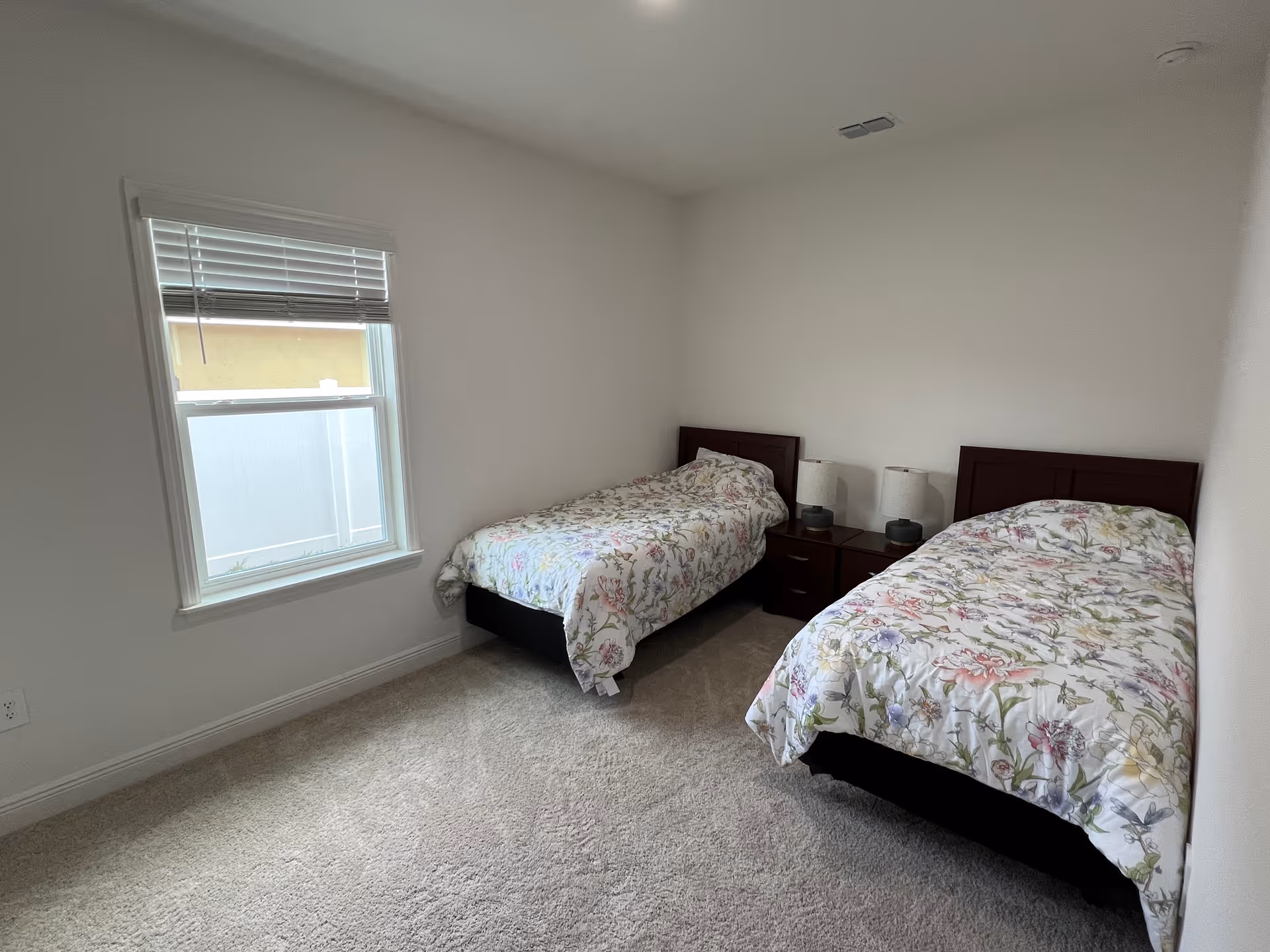 A simple bedroom with two twin beds covered in floral patterned bedspreads. Between the beds is a dark wooden nightstand with two matching lamps. There is a window with white blinds on the left wall, and the room has light-colored carpet and plain white walls.