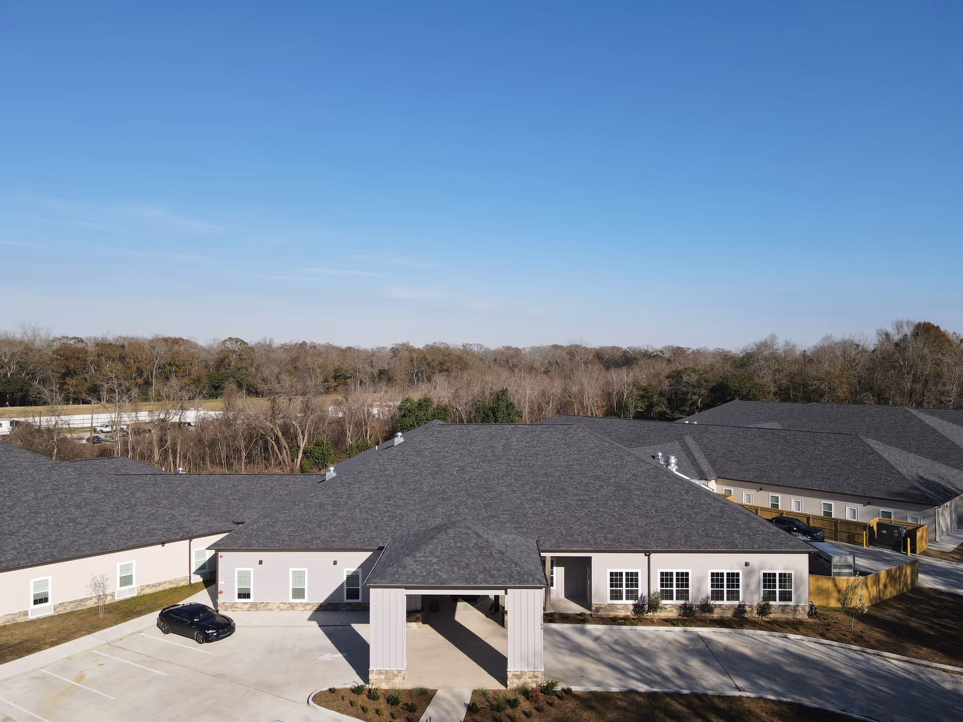 Aerial view of West Towne Assisted Living Community building with a large covered entrance, parking area with a black car, surrounded by trees under a clear blue sky.