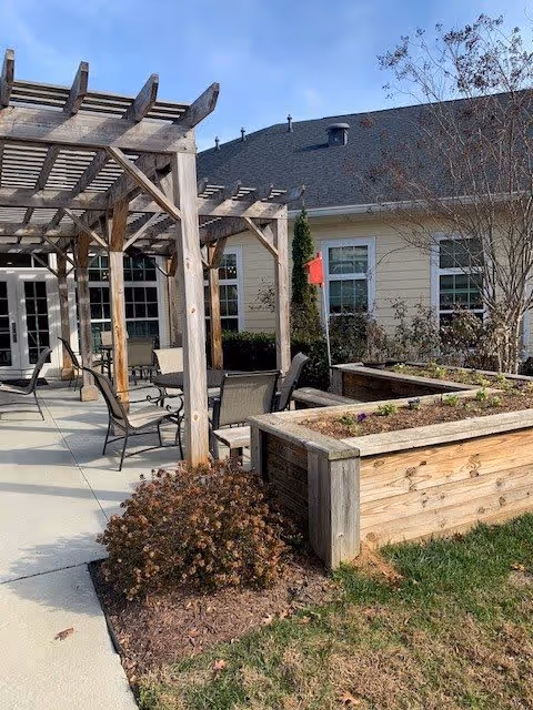 Outdoor patio area at a senior living facility featuring wooden pergolas, several chairs and tables, raised garden beds with some plants, and a building with windows in the background under a clear blue sky.