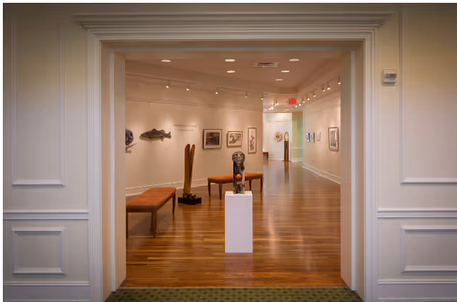 Well-lit interior gallery space with hardwood floors, sculptures on pedestals and benches, and framed artwork on the walls.