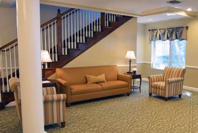Seating area with a tan sofa, two striped armchairs, side tables with lamps, and a wooden staircase in the background.