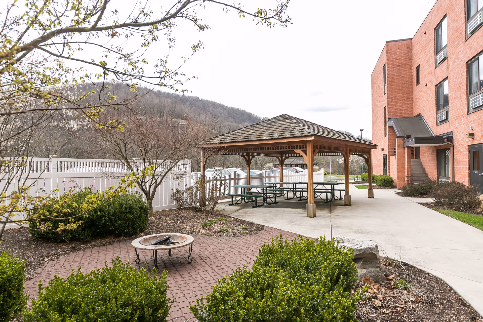 Courtyard with a wooden gazebo covering picnic tables, a small fire pit on a brick patio, surrounding shrubs and the adjacent brick building.