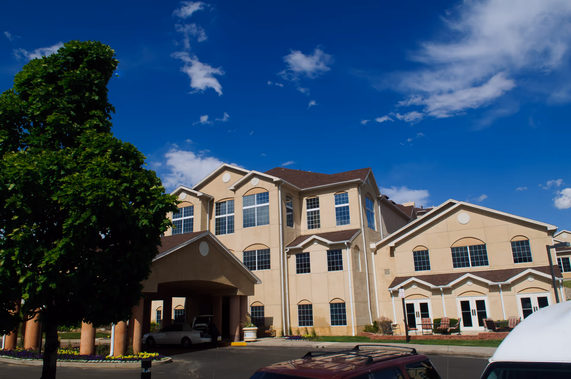 Exterior view of a multi-story beige senior living facility building under a blue sky with scattered clouds. The building has many large windows and a covered entrance with columns. There are some parked vehicles and a tree with green leaves in the foreground.