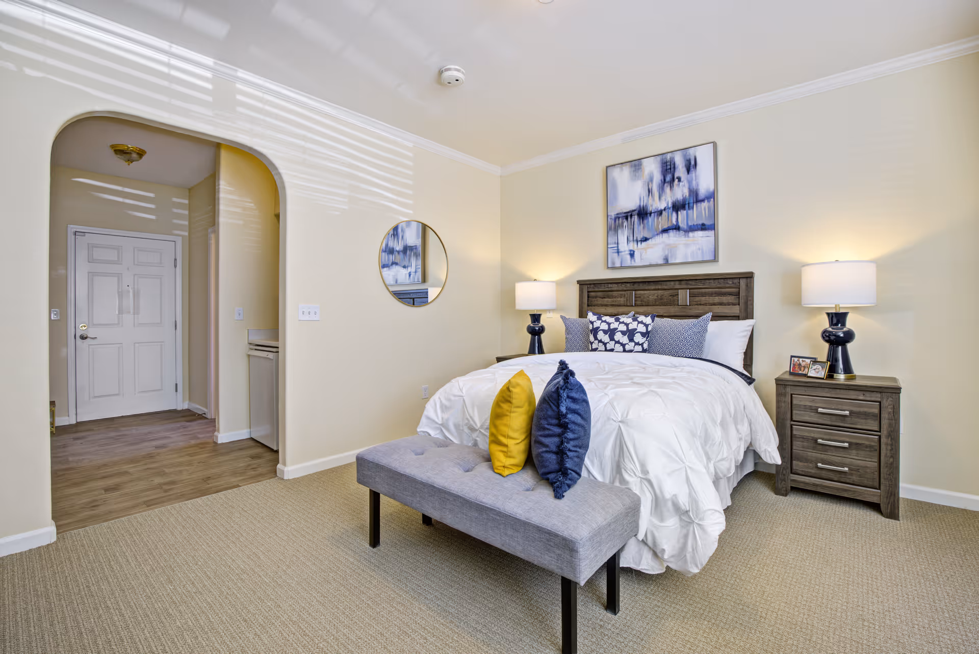 A bright and neatly arranged bedroom with a bed featuring white bedding and decorative pillows in blue and yellow. There are two matching bedside tables with lamps, a bench at the foot of the bed, a round mirror on the wall, and an abstract painting above the headboard. The room has beige walls and carpet, with an arched doorway leading to a hallway with a white door.