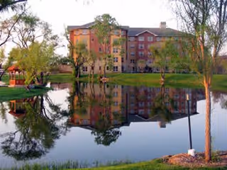 Multi-story residential building by a pond with trees and a small gazebo reflected in the water.