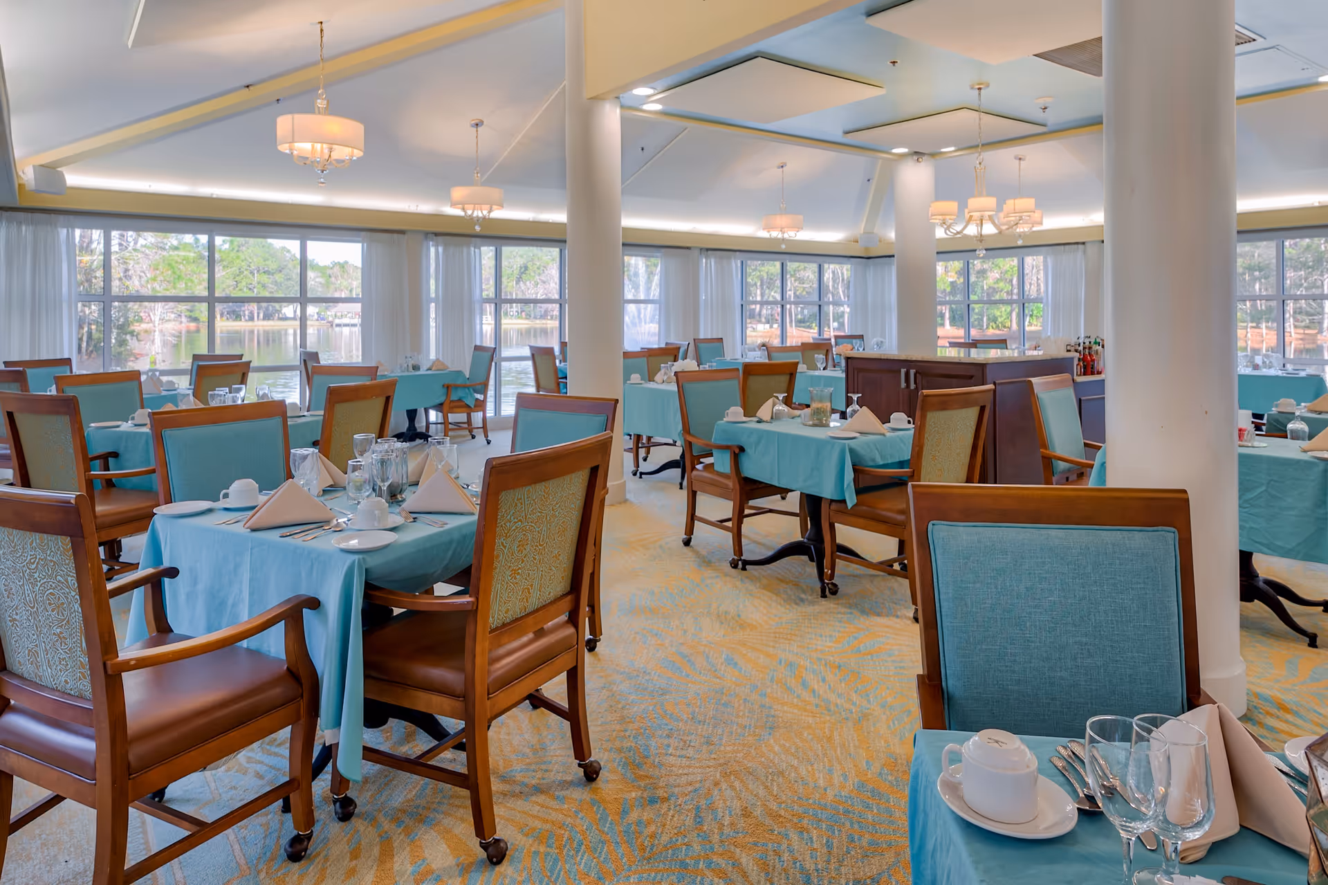 A bright dining room with large windows overlooking a lake and trees. The room features tables covered with turquoise tablecloths, each set with white napkins, plates, cups, and glassware. Wooden chairs with turquoise and patterned upholstery surround the tables. The ceiling has recessed lighting and chandeliers, and the carpet has a blue and beige pattern.