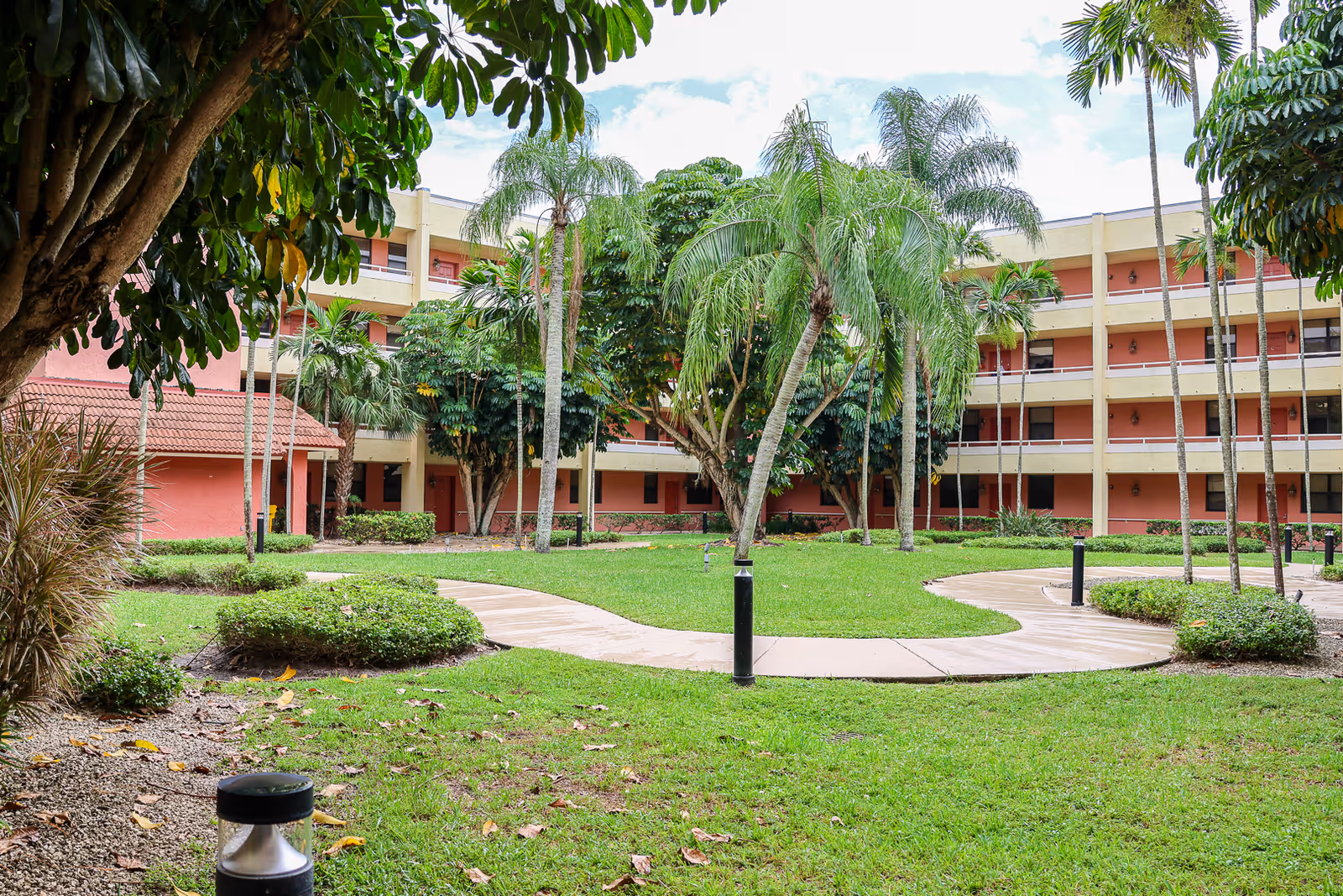 Outdoor courtyard area at Elison Independent Living of Lake Worth featuring a curved concrete walkway surrounded by green grass, palm trees, and other tropical plants. The background shows a multi-story building with balconies and a red and beige exterior.