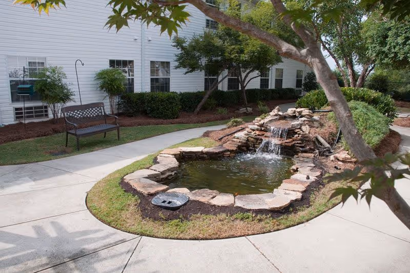 Outdoor garden area at Augusta Gardens - Senior Living & Memory Care featuring a small pond with a cascading waterfall surrounded by rocks, a concrete walkway, a bench, and greenery including bushes and trees near a white building.