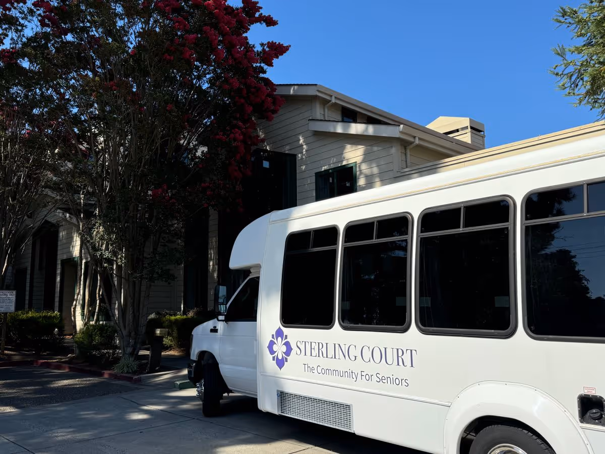 A Sterling Court senior living shuttle bus parked in front of the facility building under trees and a clear blue sky.