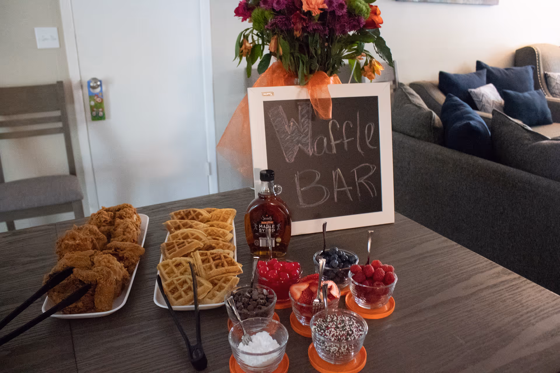 A table set up as a waffle bar with waffles, toppings, syrup and a chalkboard sign reading "Waffle BAR" in a living/dining area.