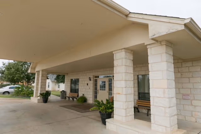 Covered stone entrance with large columns, benches, potted plants, and a doorway under a porte-cochere.