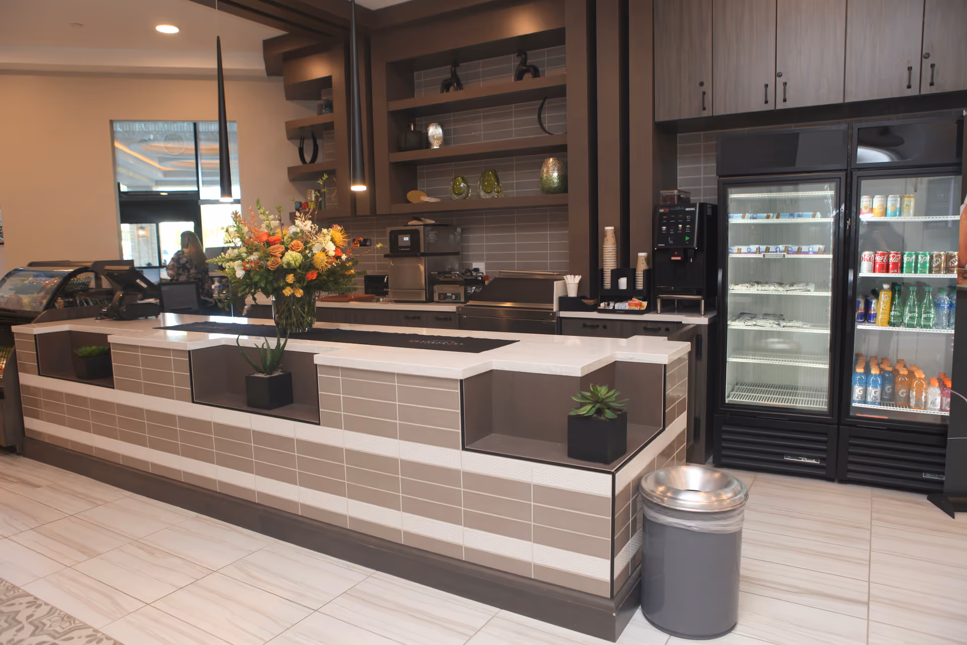 Modern café-style service counter with a flower arrangement, coffee equipment, and refrigerated beverage coolers inside a facility lobby.