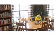 A dining area with a wooden table surrounded by wooden chairs. A vase with sunflowers and other flowers is placed in the center of the table. Large windows with checkered curtains allow natural light to fill the room. A bookshelf filled with books is visible on the left side.