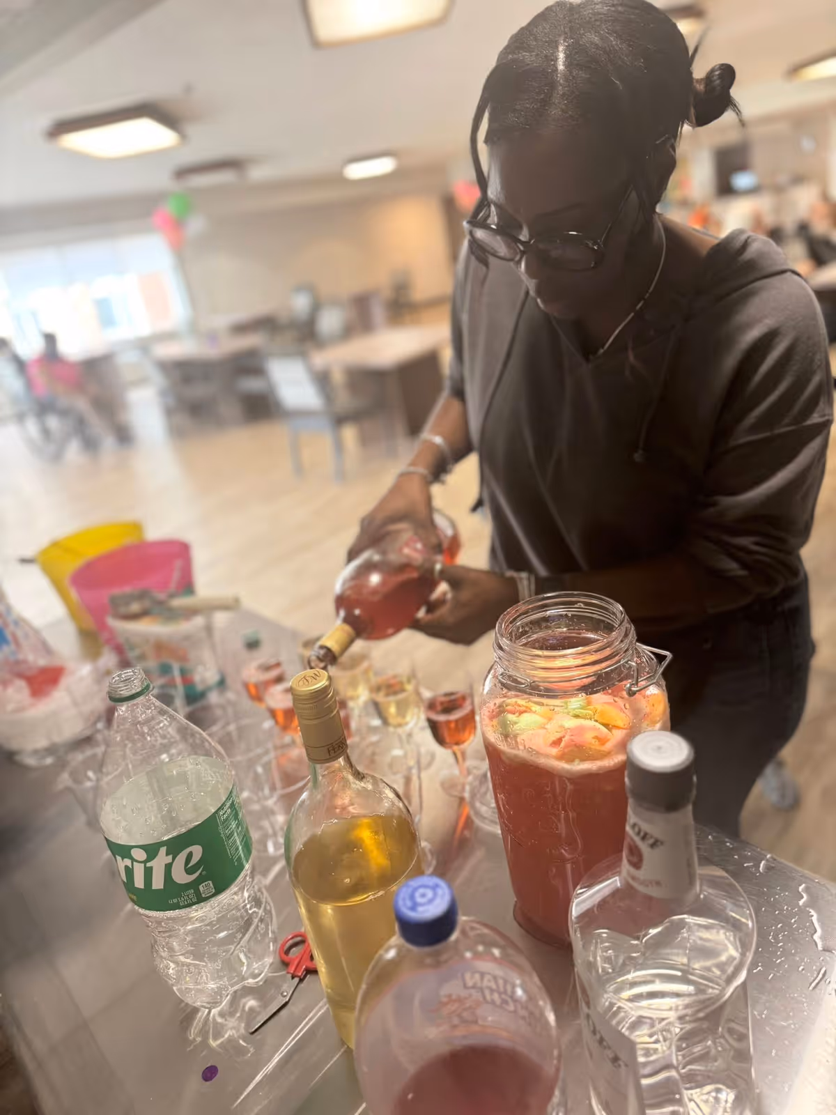 A woman wearing glasses and a dark top is pouring a pink beverage from a bottle into a glass on a table. The table has various bottles including a large bottle of Sprite, a bottle of white wine, a large jar with a fruit punch drink containing slices of fruit, and other beverage containers. The background shows a spacious room with tables and chairs, and some people sitting and moving around.