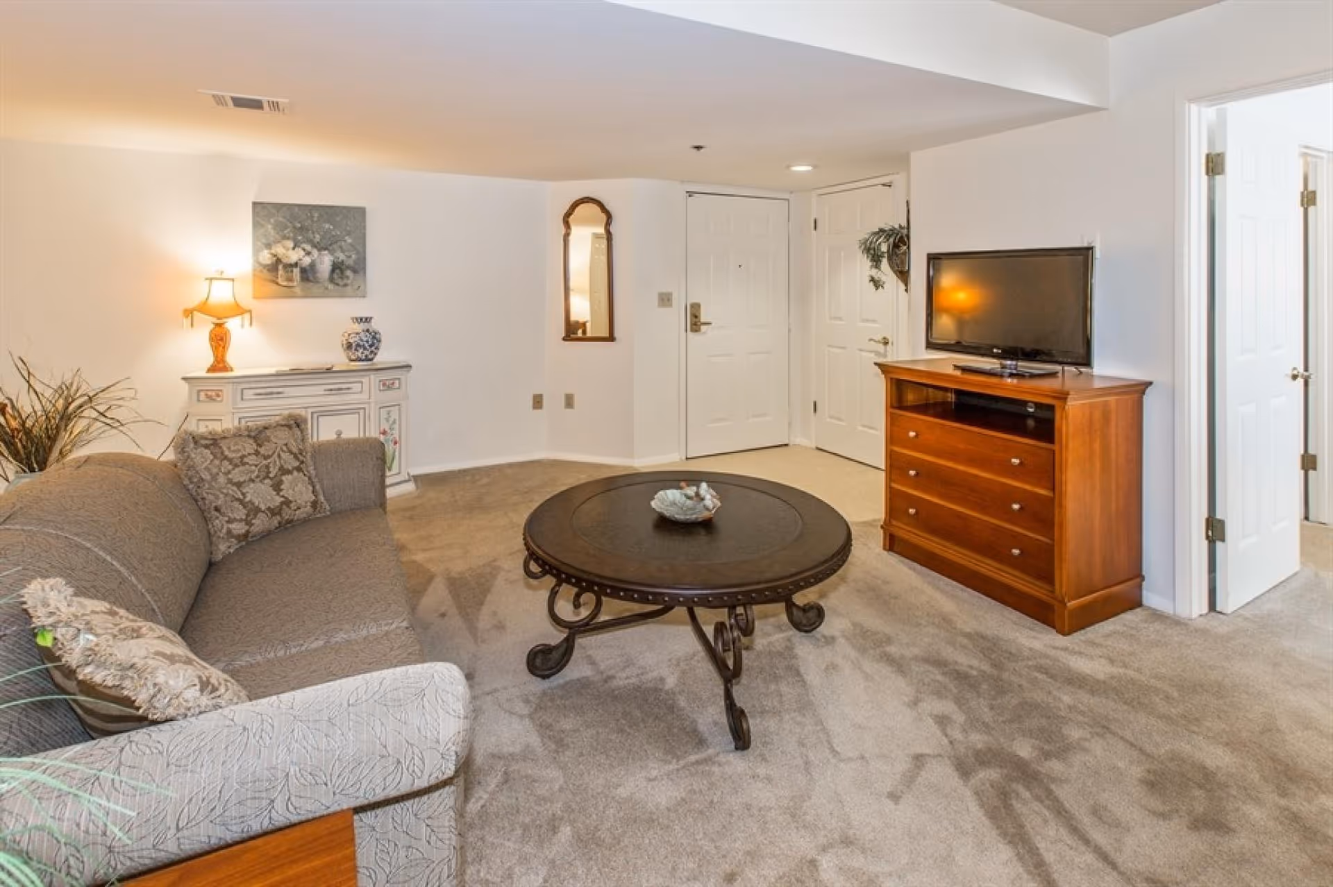 Carpeted living room with a sofa, round coffee table, TV on a wooden dresser, and an entry door in the background.