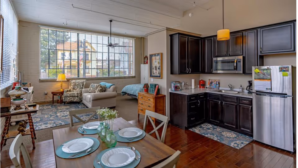 A bright, open studio apartment featuring a dining table set for four in the foreground, a kitchen with dark wood cabinets and stainless steel appliances on the right, and a cozy living area with a sofa, armchair, and a large window letting in natural light. A bed is visible in the background near the living area.